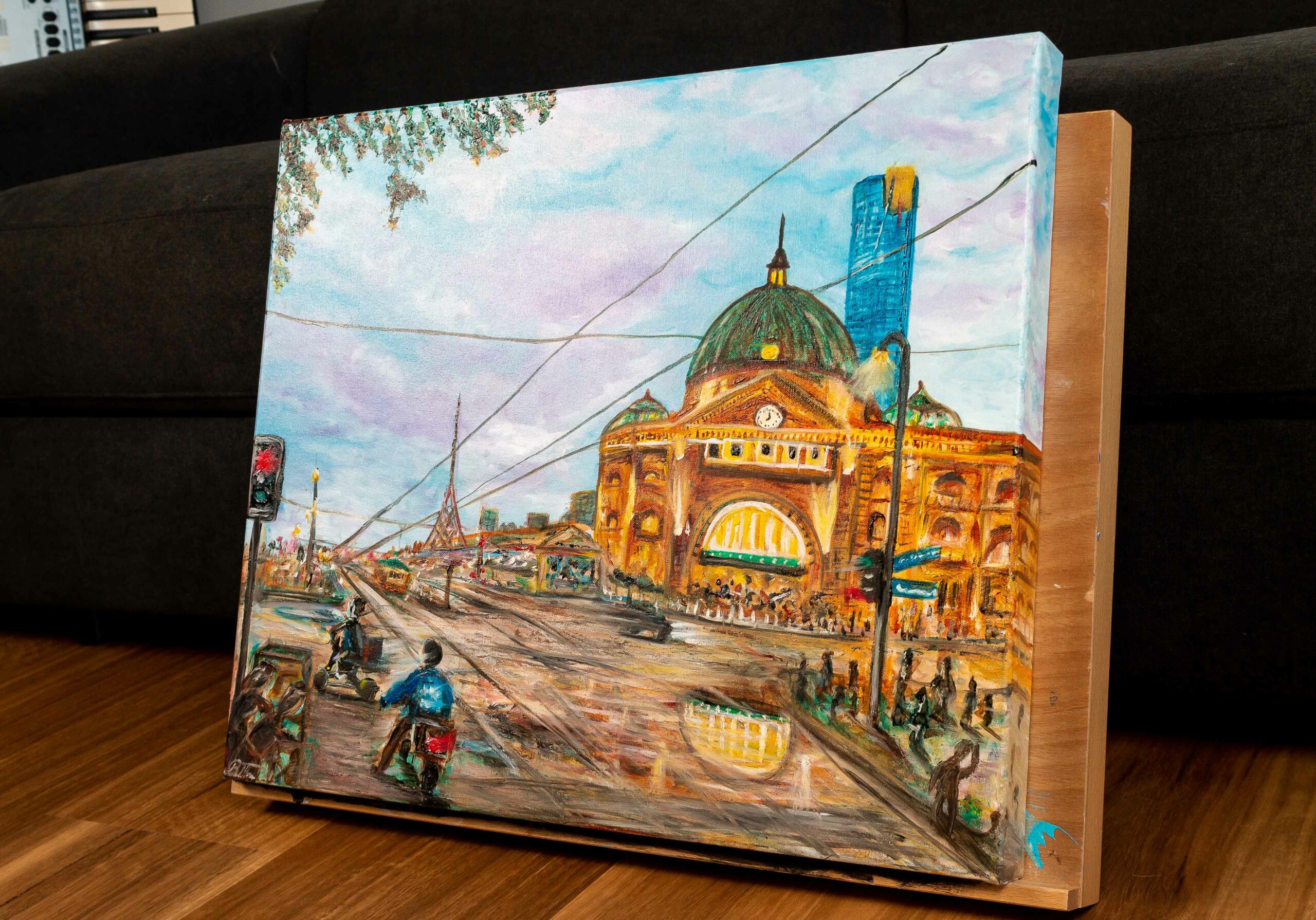Acrylic painting showing a train station lit up in yellow and gold, with a blue and purple sky, and many people/drivers/motorcyclists in the foreground going about their business.